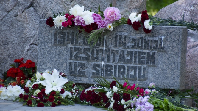 Plāksne pie piemiņas akmens ar sarkanbaltsarkaniem ziediem