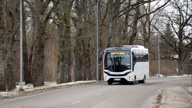 Autobuss brauc pa ceļu. Fonā pelecīga pavasara ainava ar kokiem bez lapām