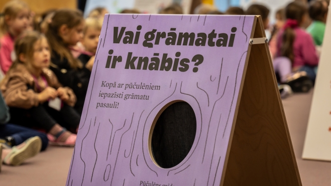Bērni bibliotēkā un lillā baneris ar tekstu "Vai grāmai ir knābis" 