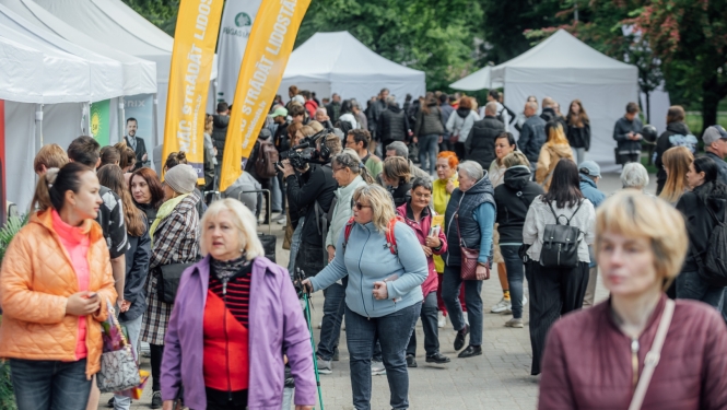 Pūlis ar cilvēkiem darba iespēju festivālā, malās baltas teltis un dzelteni vertikāli baneri