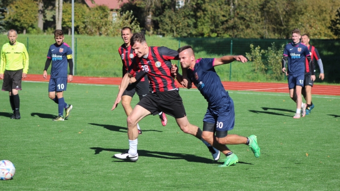 Futbolists sarkanā krekliņā cinās par futbola bumbu ar spēlētāju zilā kreklā