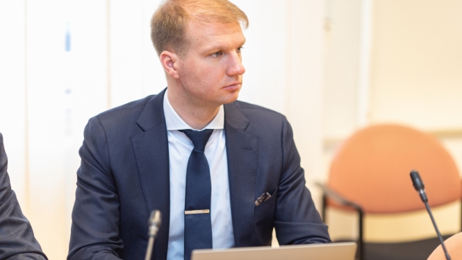 Vīrietis ar gaišiem matiem tumši zilā uzvalkā ar kaklasaiti sēž pie sarunu galda