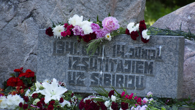 Plāksne pie piemiņas akmens ar sarkanbaltsarkaniem ziediem