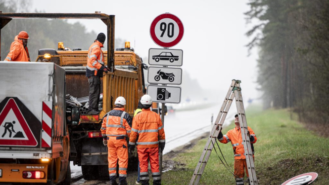 Ceļa strādnieki oranžos tērpos liek ceļa zīmes