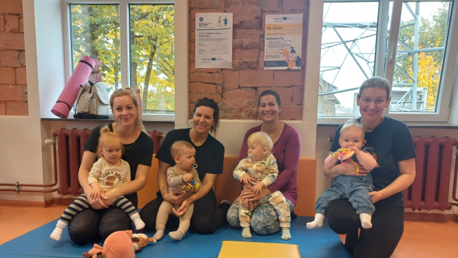 4 smaidīgas māmiņas ar maziem bērniem klēpjos uz vingrošanas paklāja