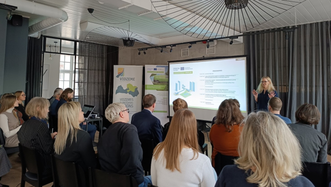 Cilvēki sēž zālē un klausās semināra prezentāciju