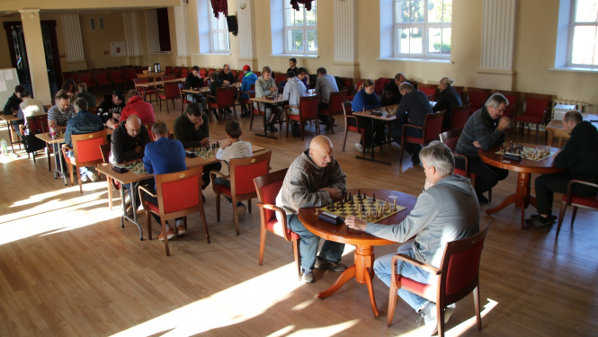 Turnīra dalībnieki kultūras nama zālē sēž pie galdiem un spēlē šahu