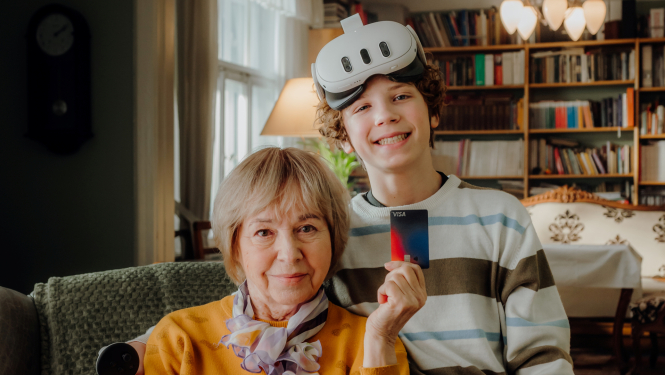 Dāma gados ar īsiem gaišiem matiem tur rokā norēķinu karti un smaidīgs jauneklis ar VR brillēm