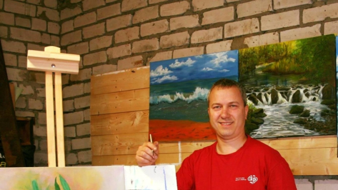 Vīrietis sarkanā kreklā pie molberta ar gleznu