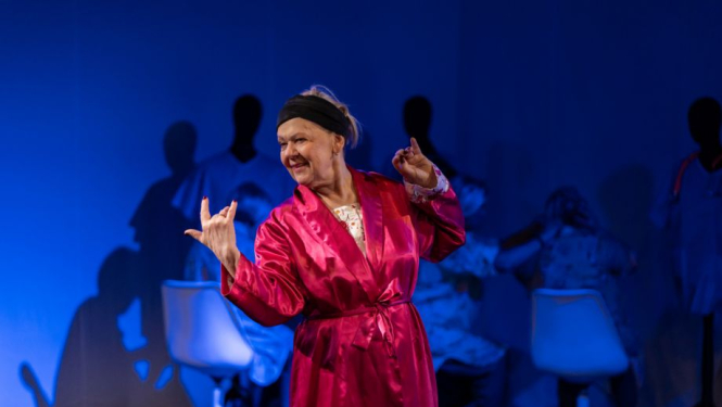 Aktieris sarkanā tērpā uz zila skatuves fona