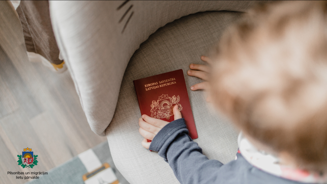 Mājīga iekštelpu aina, kurā redzama bērna roka, kas sniedzas pēc sarkanas Latvijas pases, kas atrodas uz mīksta, gaiša krēsla.