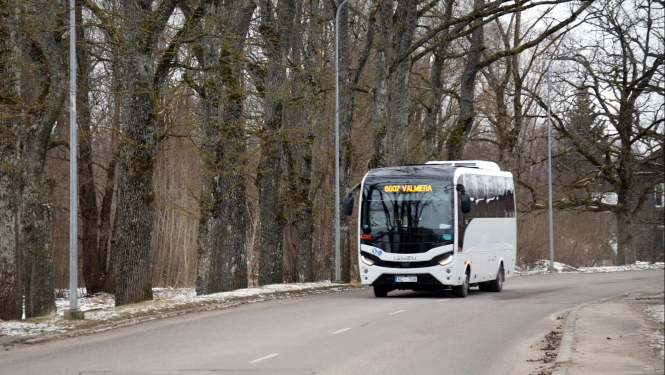Autobuss brauc pa ceļu. Fonā pelecīga pavasara ainava ar kokiem bez lapām