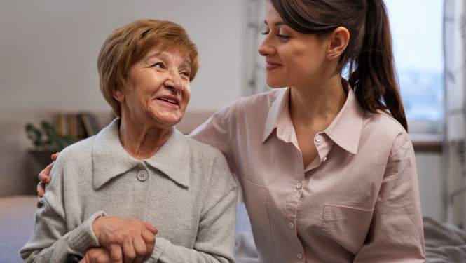 Smaidīga sieviete apķērusi ap plecu smaidīgu kundzi gados