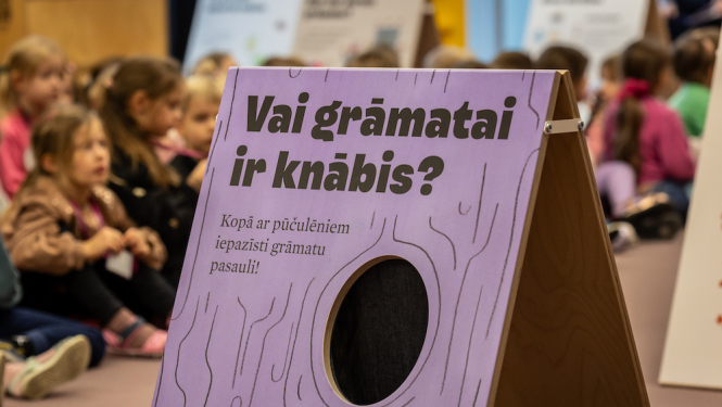 Bērni bibliotēkā un lillā baneris ar tekstu "Vai grāmai ir knābis" 