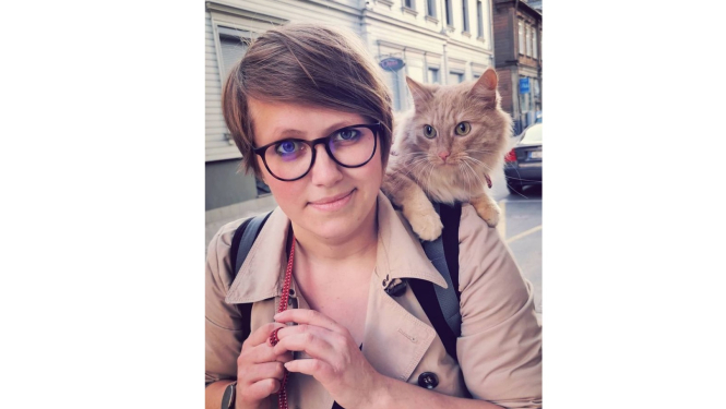 Sieviete ar īsiem, gaiši brūniem matiem. Viņai uz pleca ruds kaķis.