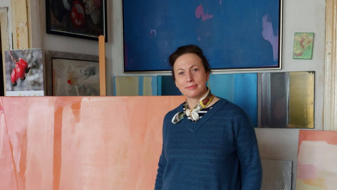 Sieviete ar tumšiem matiem zilā džemperī stāv pie gleznām