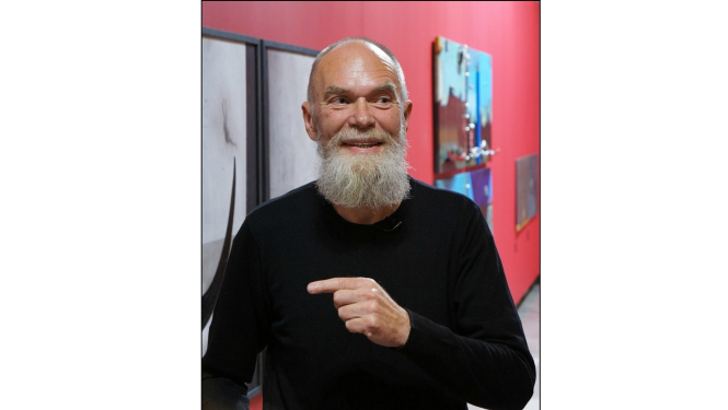 Portrets. Smaidošs pusmūža vīrietis ar baltu bārdu un gleznām fonā