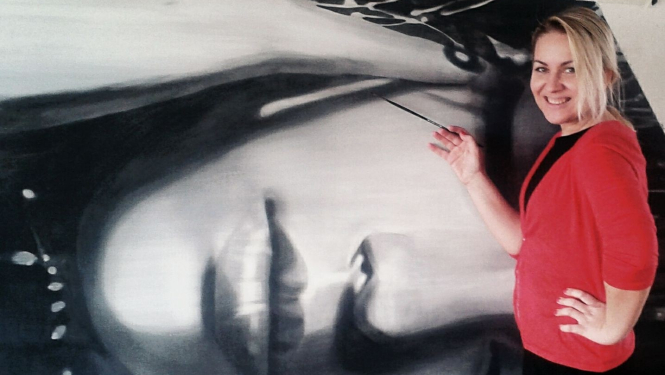 Māksliniece ar gaišiem matiem, sarkanā žaketē ar otu rokās pie lielizmērā mākslas darbu, kurā atspoguļots sievietes portrets