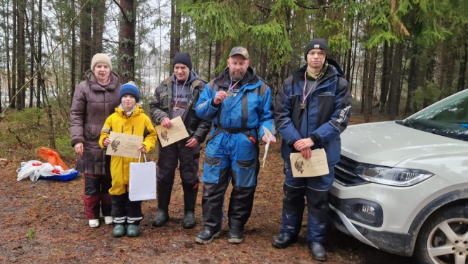 Kopbilde ar sacensību uzvarētājiem - viena sieviete, viens bērns dzeltenā jaciņā, trīs pieauguši vīrieši ar diplomiem un medaļām.
