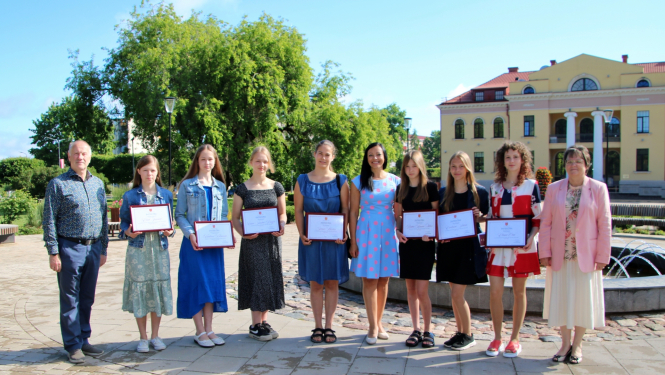 Cilvēki pie strūklakas, fonā koks ar zaļām lapām un dzeltena kultūras nama ēka
