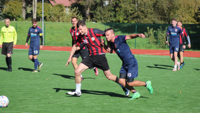 Futbolists sarkanā krekliņā cinās par futbola bumbu ar spēlētāju zilā kreklā