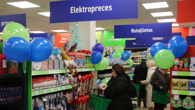 Rosīgā veikalā pircēji pārlūko labi aprīkotu eju, kas apzīmēta ar “Elektropreces”. Telpu rotā krāsaini baloni, kas kontrastē ar plauktiem, kas piepildīti ar dažādiem produktiem, tostarp rotaļlietām, mājas precēm un dekorācijām.