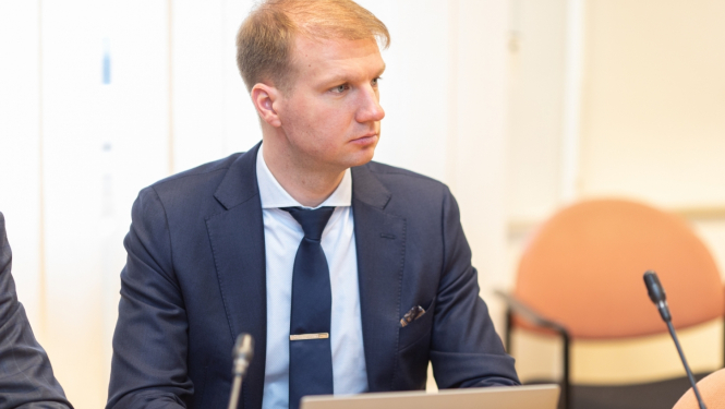 Vīrietis ar gaišiem matiem tumši zilā uzvalkā ar kaklasaiti sēž pie sarunu galda