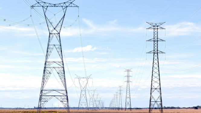 Plaša, atklāta ainava zem skaidrām zilām debesīm, kas izraibinātas ar augstām, metāliskām elektropārvades līnijām un piloniem, kas stiepjas tālumā. Priekšplānā ir zeltains zāles lauks.