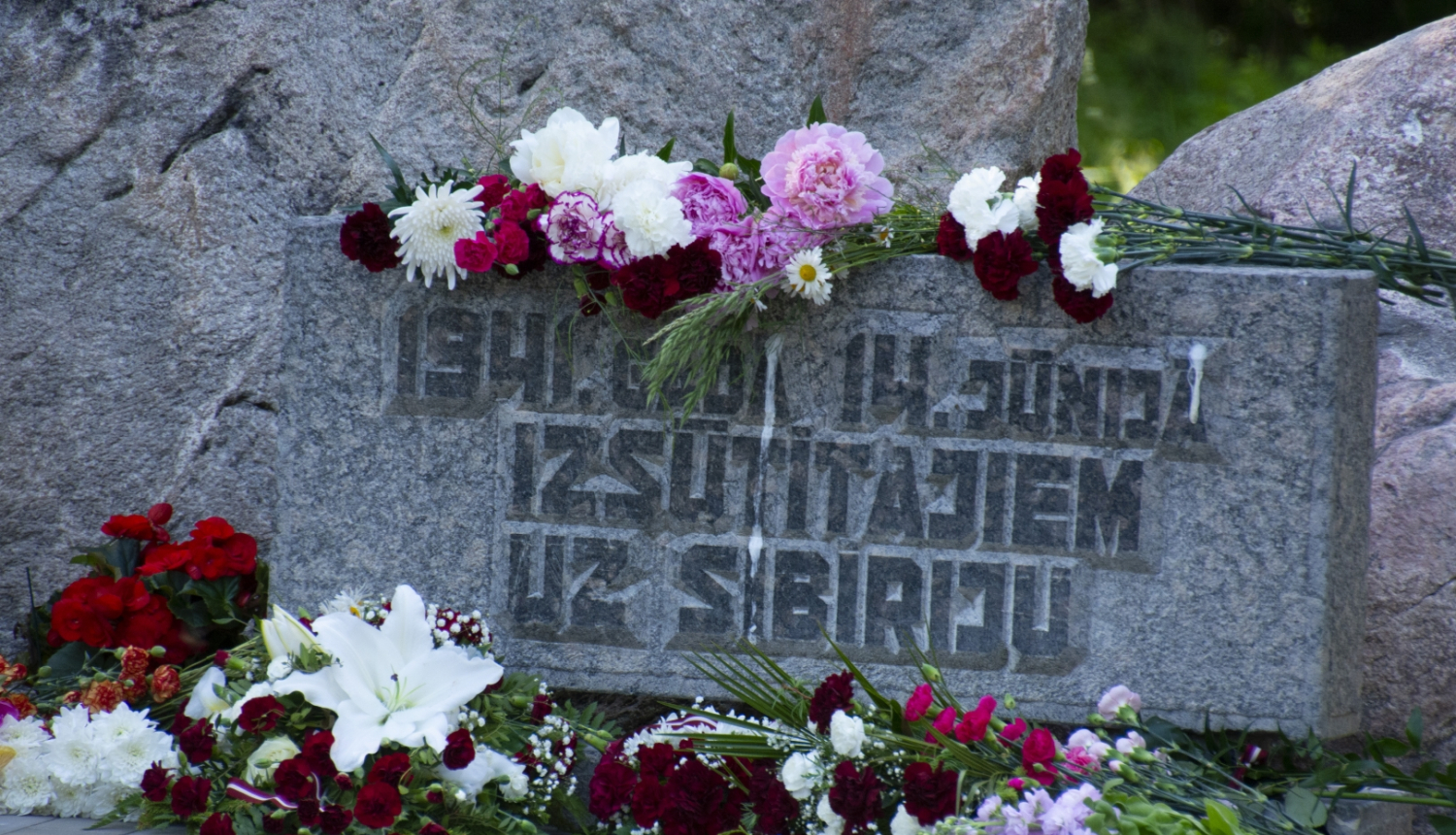 Plāksne pie piemiņas akmens ar sarkanbaltsarkaniem ziediem