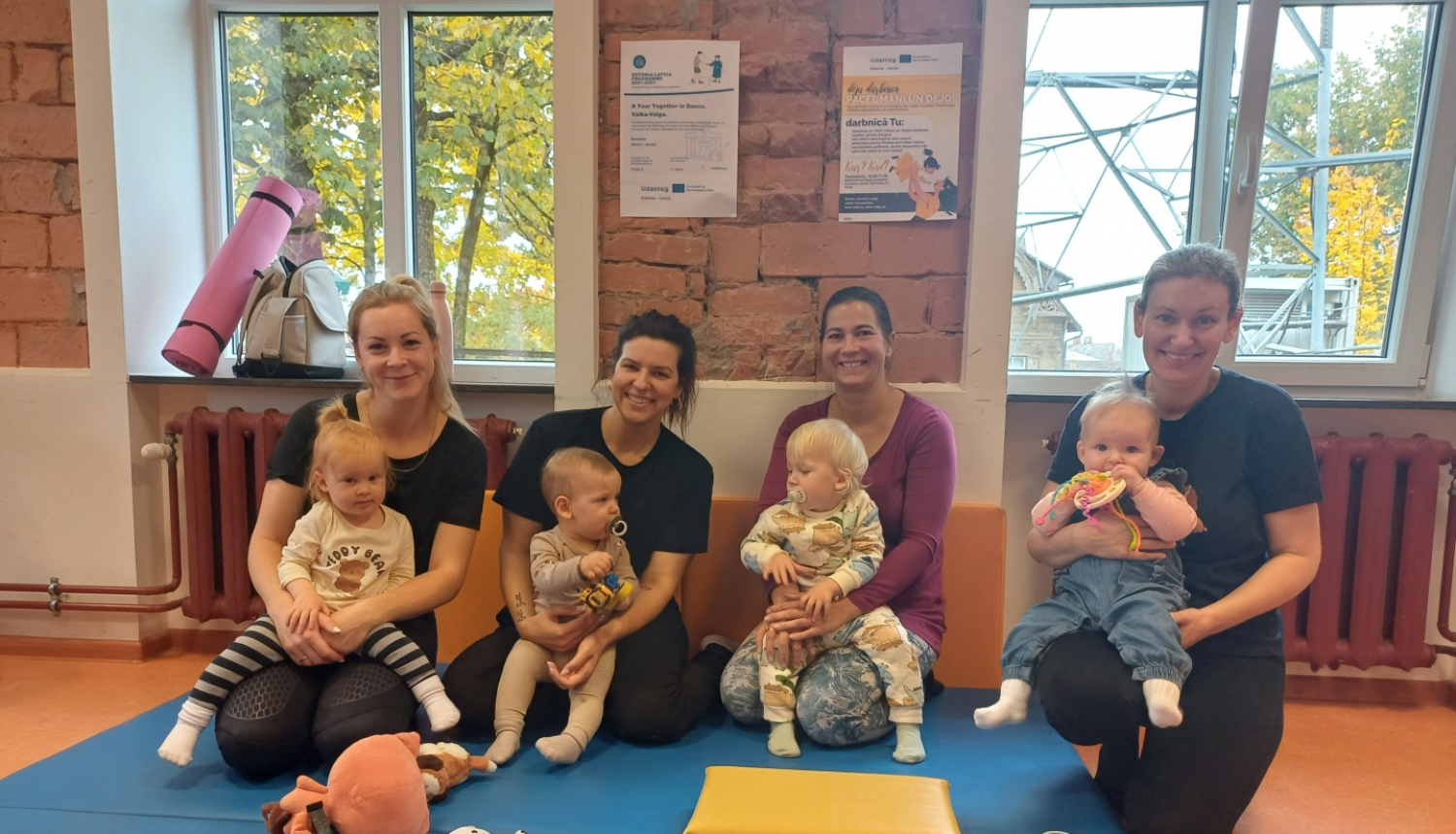 4 smaidīgas māmiņas ar maziem bērniem klēpjos uz vingrošanas paklāja