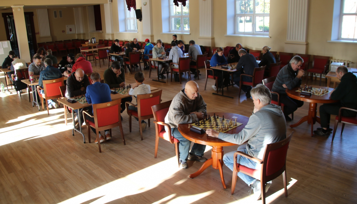 Turnīra dalībnieki kultūras nama zālē sēž pie galdiem un spēlē šahu
