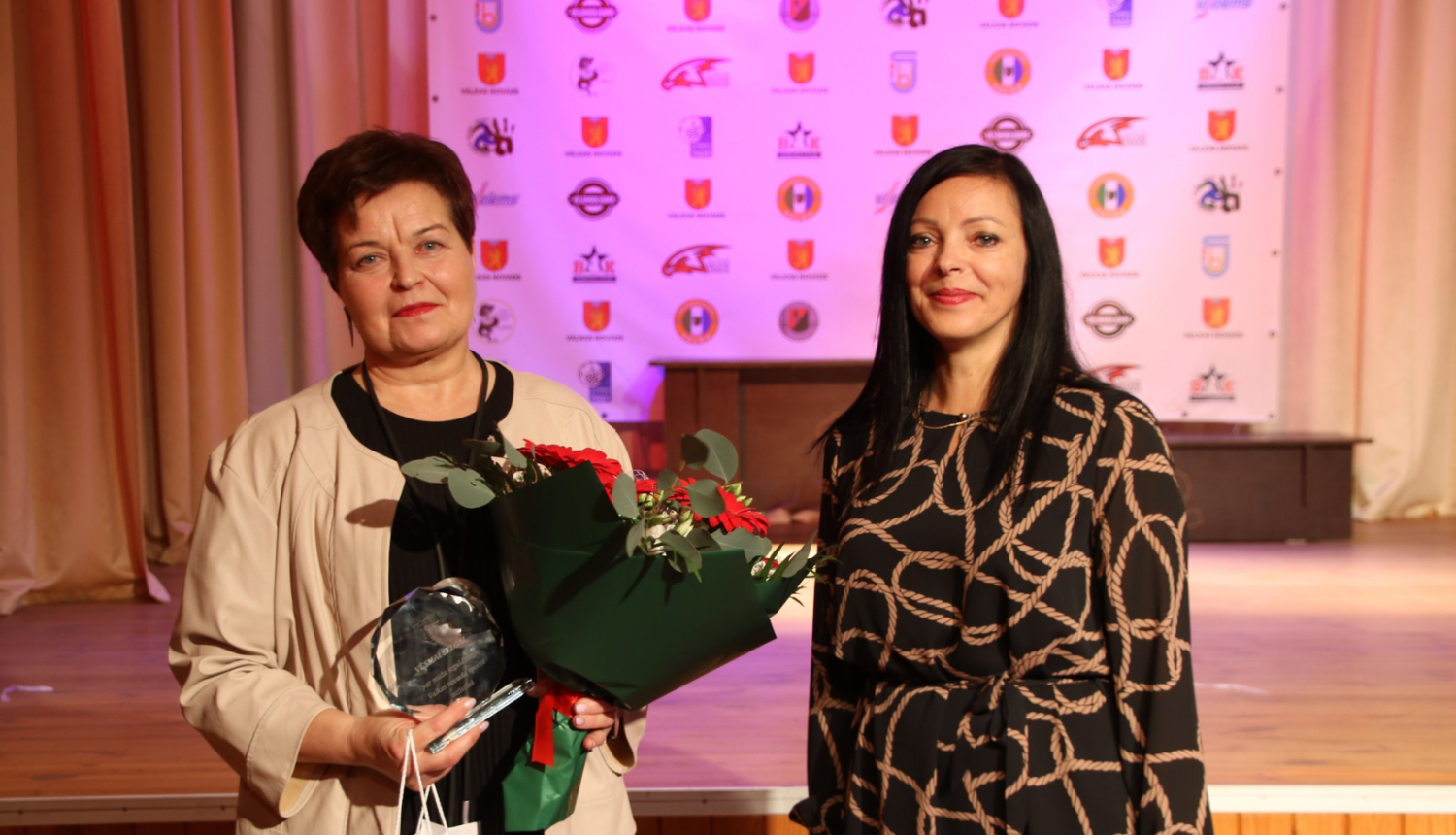 Divas sievietes ar turmšiem matiem, vienai rokās ziedu pušķi. 