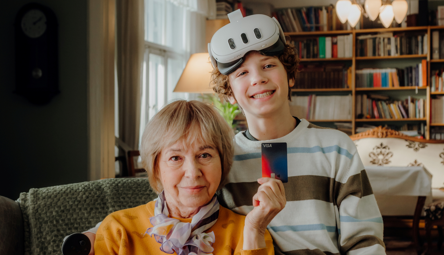 Dāma gados ar īsiem gaišiem matiem tur rokā norēķinu karti un smaidīgs jauneklis ar VR brillēm