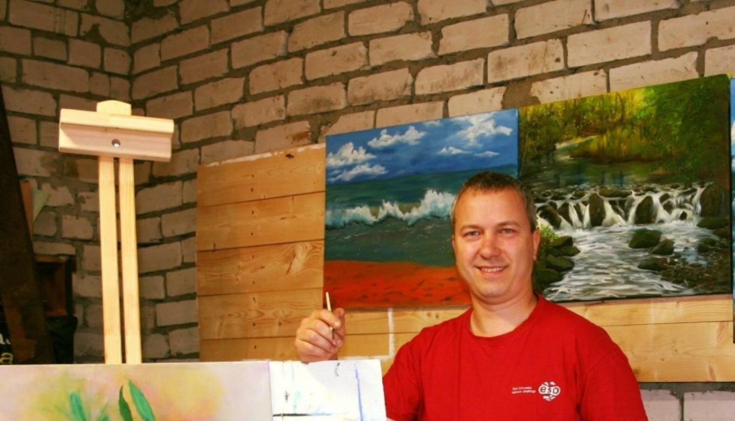 Vīrietis sarkanā kreklā pie molberta ar gleznu