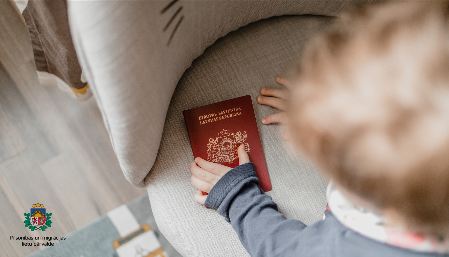 Mājīga iekštelpu aina, kurā redzama bērna roka, kas sniedzas pēc sarkanas Latvijas pases, kas atrodas uz mīksta, gaiša krēsla.