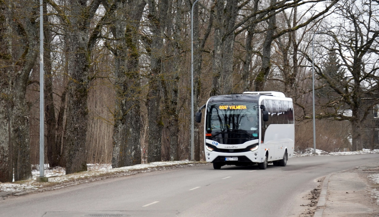 Autobuss brauc pa ceļu. Fonā pelecīga pavasara ainava ar kokiem bez lapām