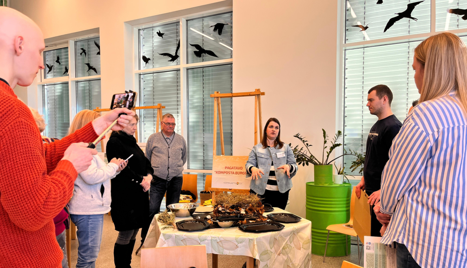 Cilvēki sastājušies ap galdu klausās stāstījumu par kompostiem