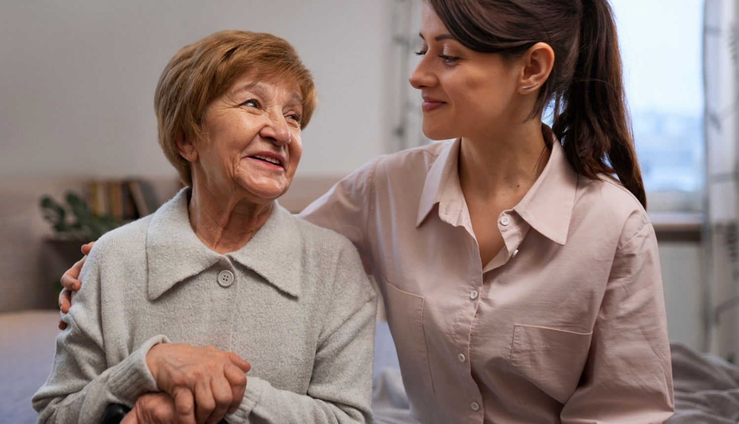 Smaidīga sieviete apķērusi ap plecu smaidīgu kundzi gados