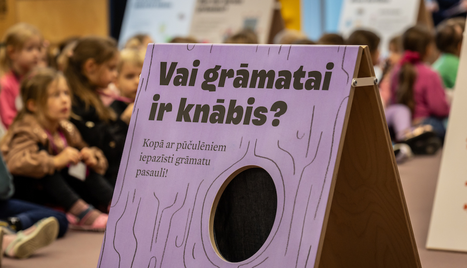 Bērni bibliotēkā un lillā baneris ar tekstu "Vai grāmai ir knābis" 