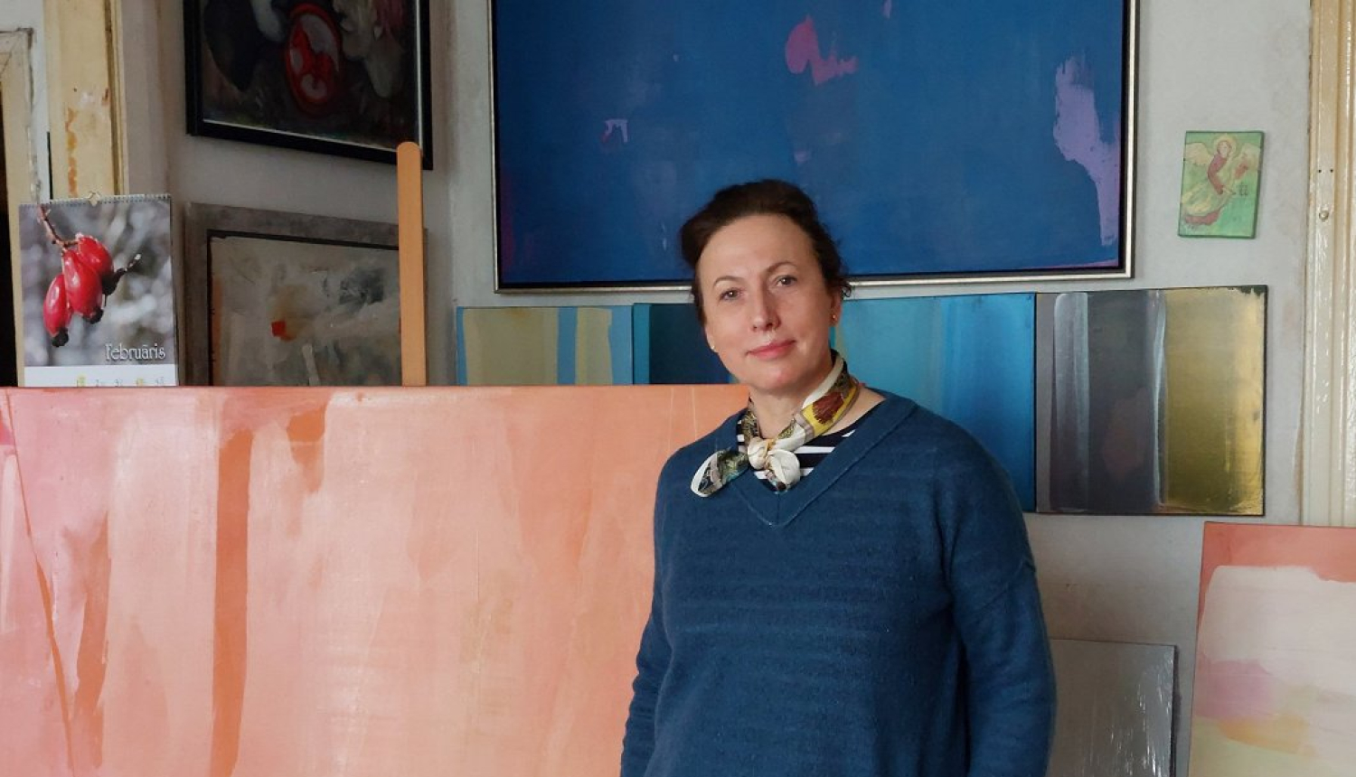 Sieviete ar tumšiem matiem zilā džemperī stāv pie gleznām