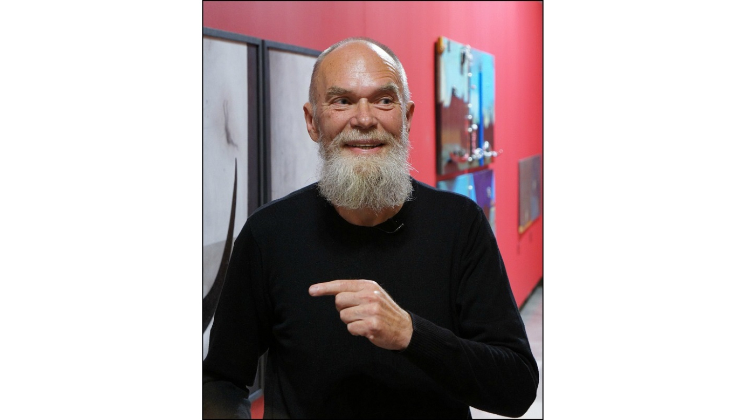 Portrets. Smaidošs pusmūža vīrietis ar baltu bārdu un gleznām fonā