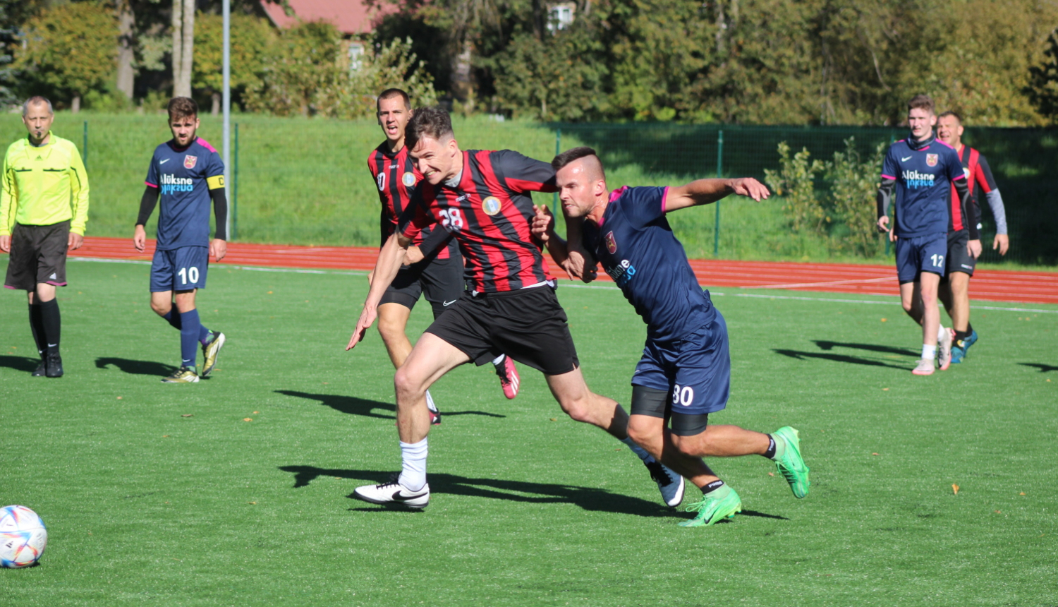 Futbolists sarkanā krekliņā cinās par futbola bumbu ar spēlētāju zilā kreklā