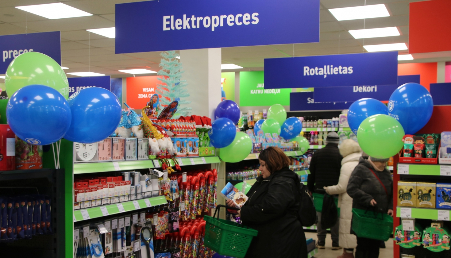 Rosīgā veikalā pircēji pārlūko labi aprīkotu eju, kas apzīmēta ar “Elektropreces”. Telpu rotā krāsaini baloni, kas kontrastē ar plauktiem, kas piepildīti ar dažādiem produktiem, tostarp rotaļlietām, mājas precēm un dekorācijām.