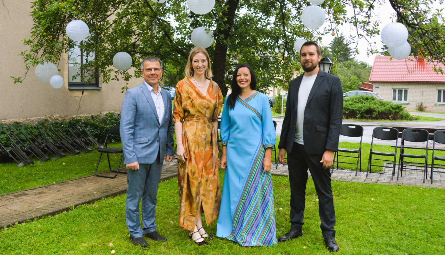 Divi vīrieši uzvalkos un divas sievietes kleitās muzeja pagalmā pie koka ar balonu dekorācijām