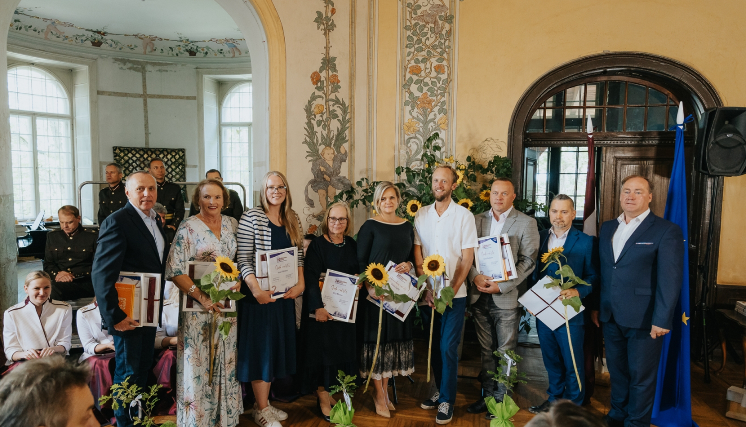 Cilvēkie svinīgā apģērbā pie Eiropas Savienības karoga