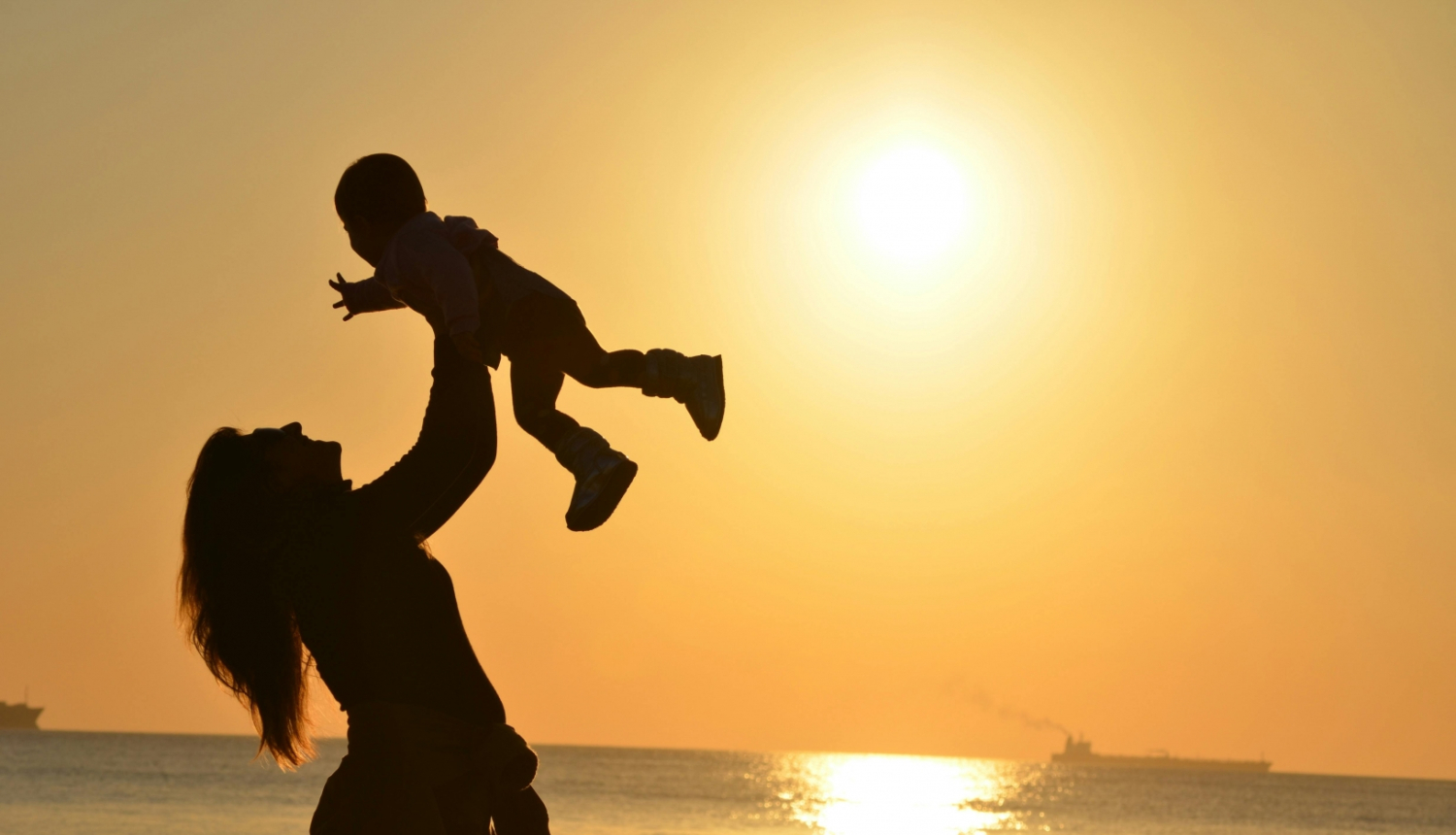 Sieviete pacēlusi rokās bērnu saulrietā