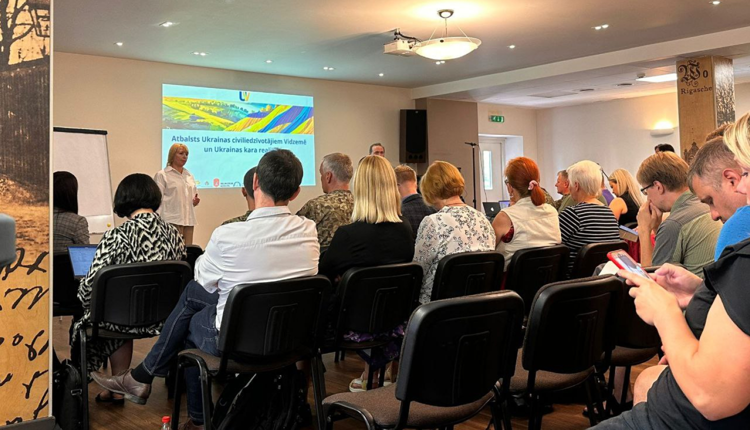 Cilvēki sēž krēslos un klausās lekciju