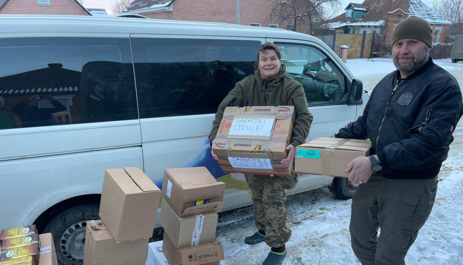 Vīrietis un sieviete pie balta busiņa ar ziedomjumu kastēm