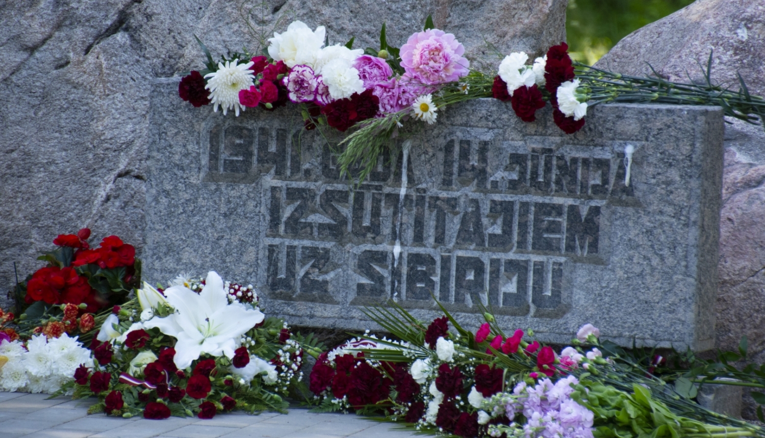 Plāksne pie piemiņas akmens ar sarkanbaltsarkaniem ziediem