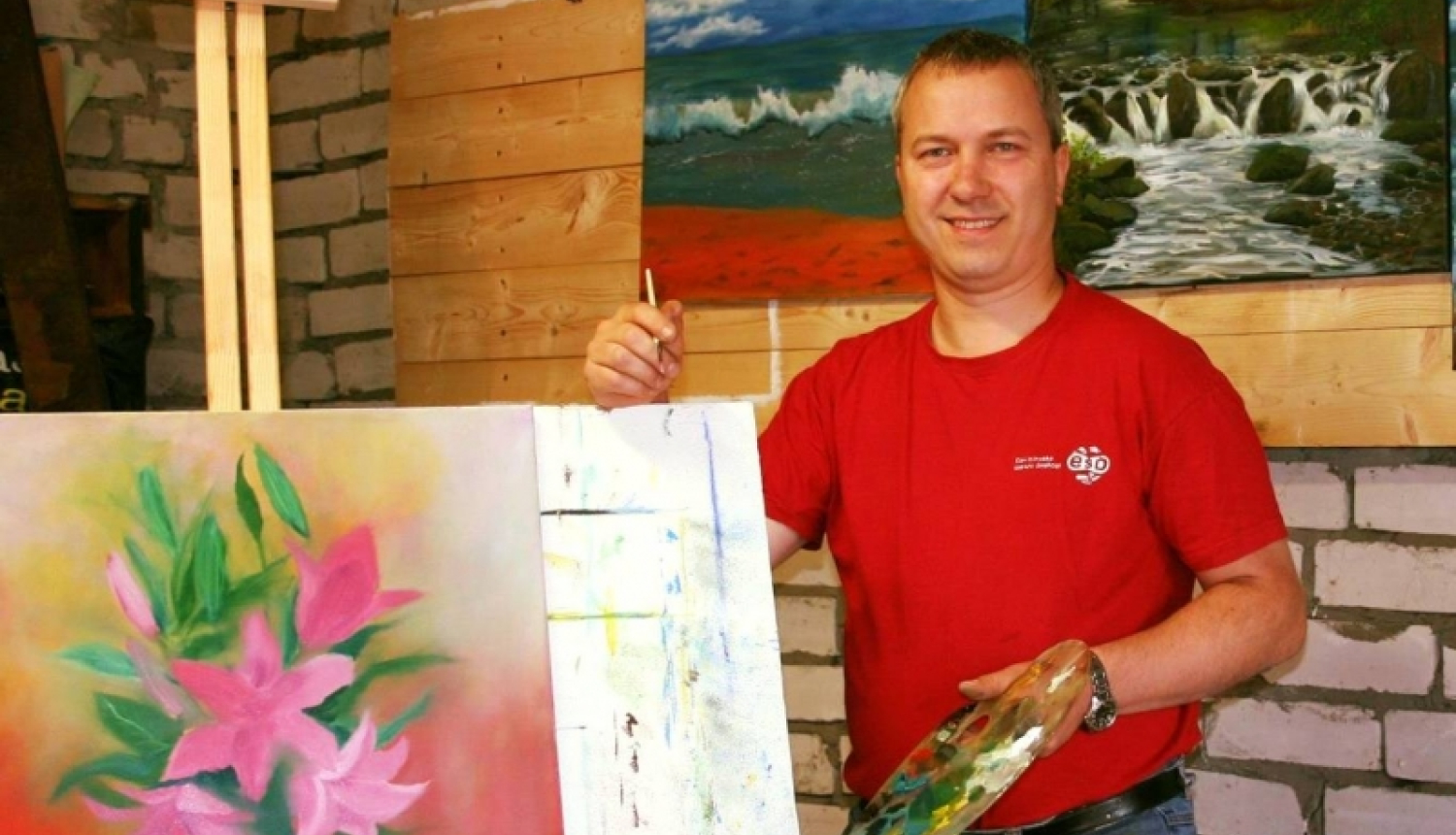 Vīrietis sarkanā kreklā pie molberta ar gleznu